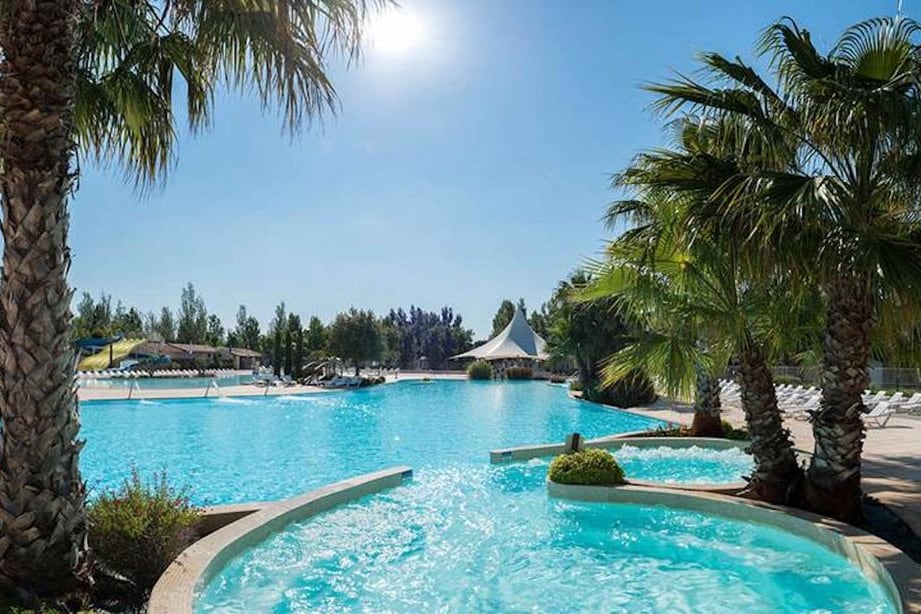 A large outdoor pool area is surrounded by palm trees, with clear blue water reflecting sunlight. Loungers are positioned around the pool, and a white tent structure is visible in the background, providing shade. The vibrant greenery enhances the relaxing atmosphere.