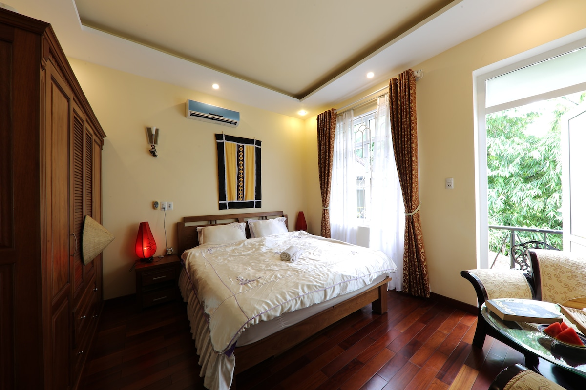 A spacious room features a large bed, complemented by wooden flooring and well-preserved wooden furniture. Natural light floods in through a window, which opens to a balcony. Decorative curtains frame the entrance, and two accent lamps add a warm touch to the atmosphere.
