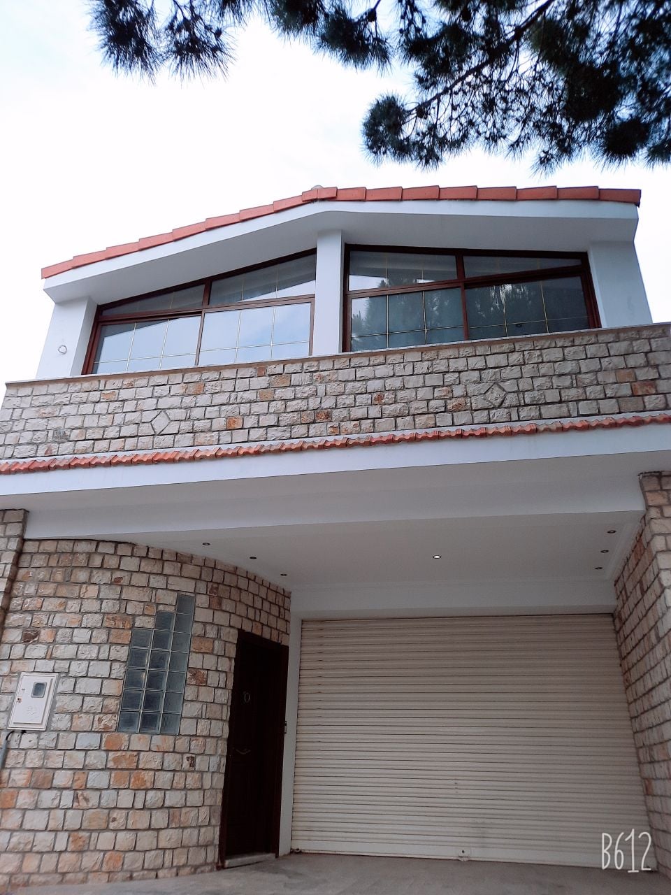 The exterior of the house is characterized by a stone façade and large windows that allow natural light to enter. A gently sloping roof complements the modern design. A secured garage door is visible, providing convenient parking for vehicles.