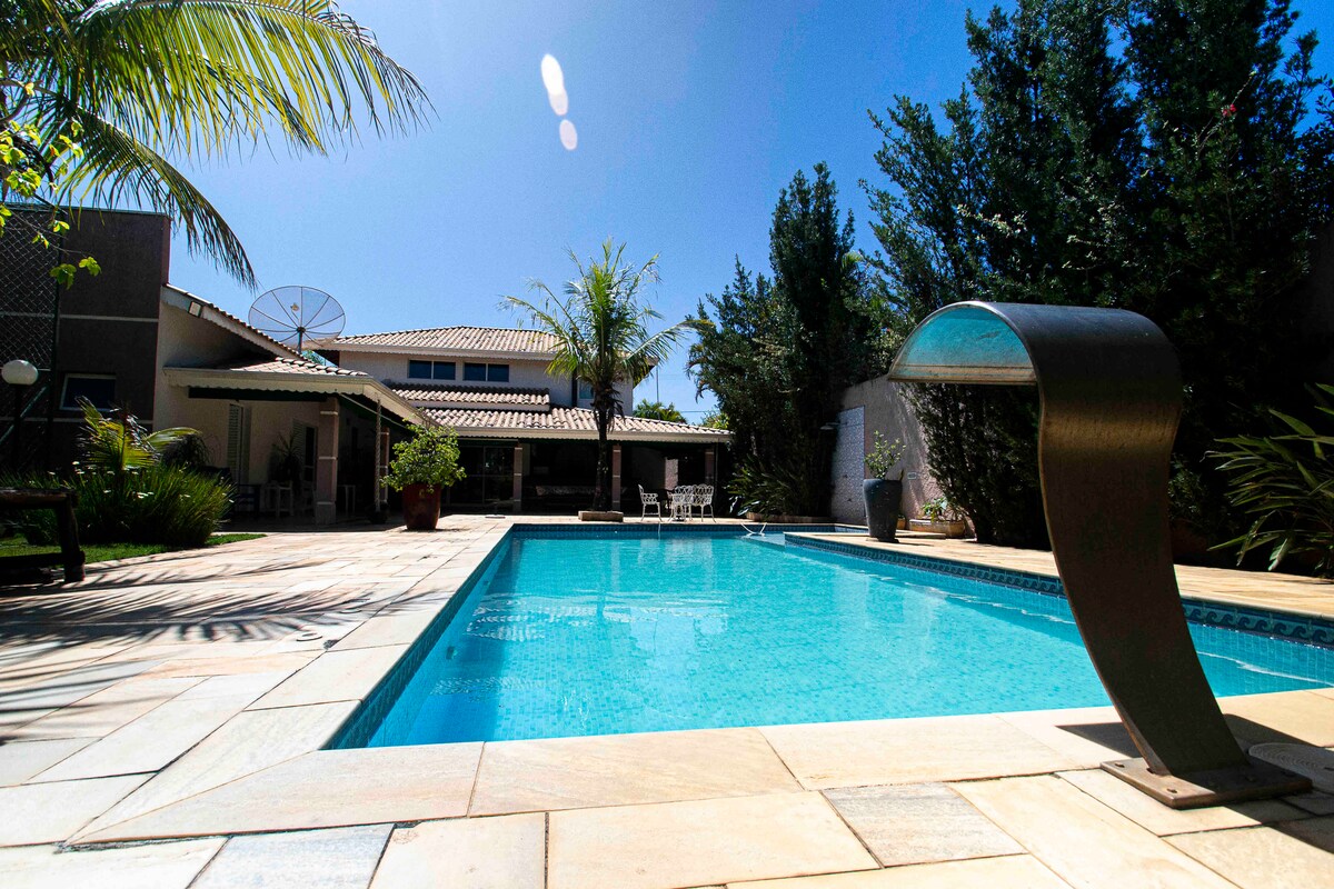 A private swimming pool is framed by a sunlit patio, featuring smooth stone tiles and surrounded by lush greenery. Palm trees provide shade, and a modern water feature adds a sleek touch. The clear blue water reflects the clear sky, creating a serene outdoor setting.