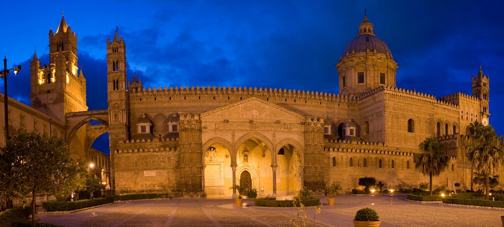 The image showcases the majestic façade of Palermo Cathedral under a twilight sky. Its intricate details, including arches and towers, are highlighted, creating a striking silhouette. Lush greenery and illuminated pathways lead toward the building, enhancing the historical significance of this UNESCO World Heritage site.