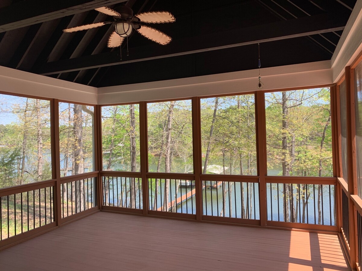 A screened porch features large windows offering views of the lake and surrounding trees. The porch is designed with wood railings and light-colored flooring, enhancing the open, airy feel. Ceiling fan and beams accentuate the rustic charm of the space.