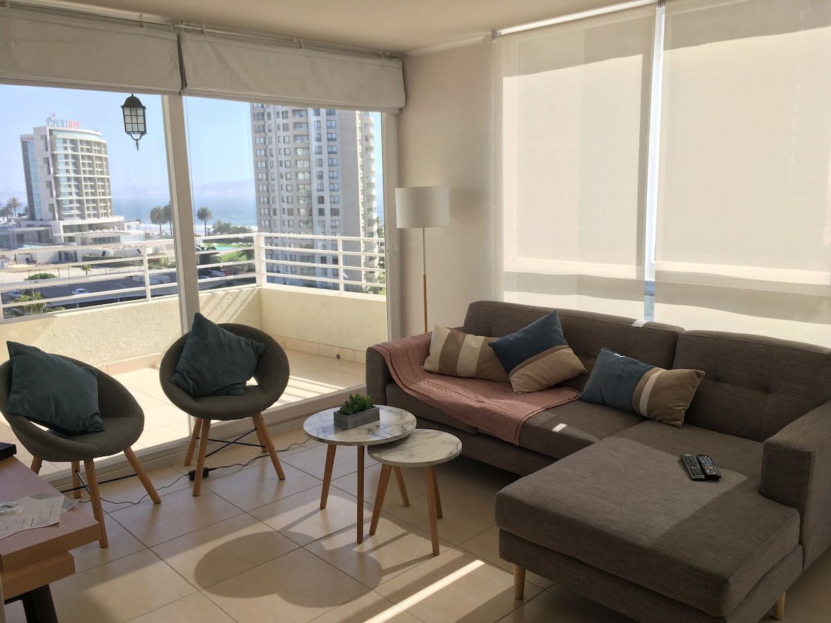 A bright living area is presented with large windows offering city views. Comfortable seating includes a spacious sectional sofa adorned with cushions, along with two accent chairs. A small coffee table with a decorative plant is situated in front, enhancing the inviting layout.