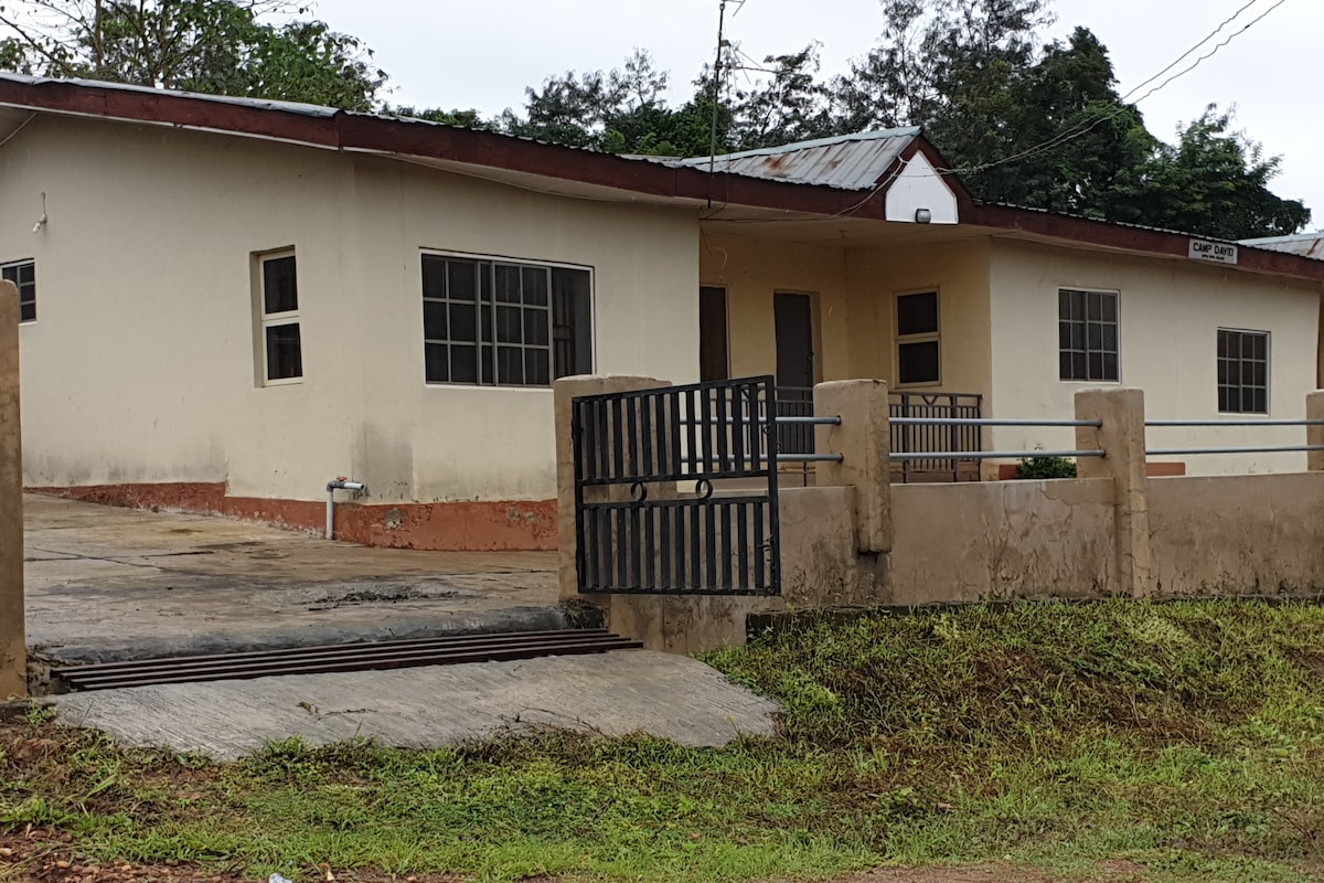 A single-story building is shown, featuring a wide concrete entrance and a gated driveway. The exterior walls are painted in light colors, with several windows providing natural light. Surrounding greenery adds to the tranquil setting, creating an inviting approach to the facility.