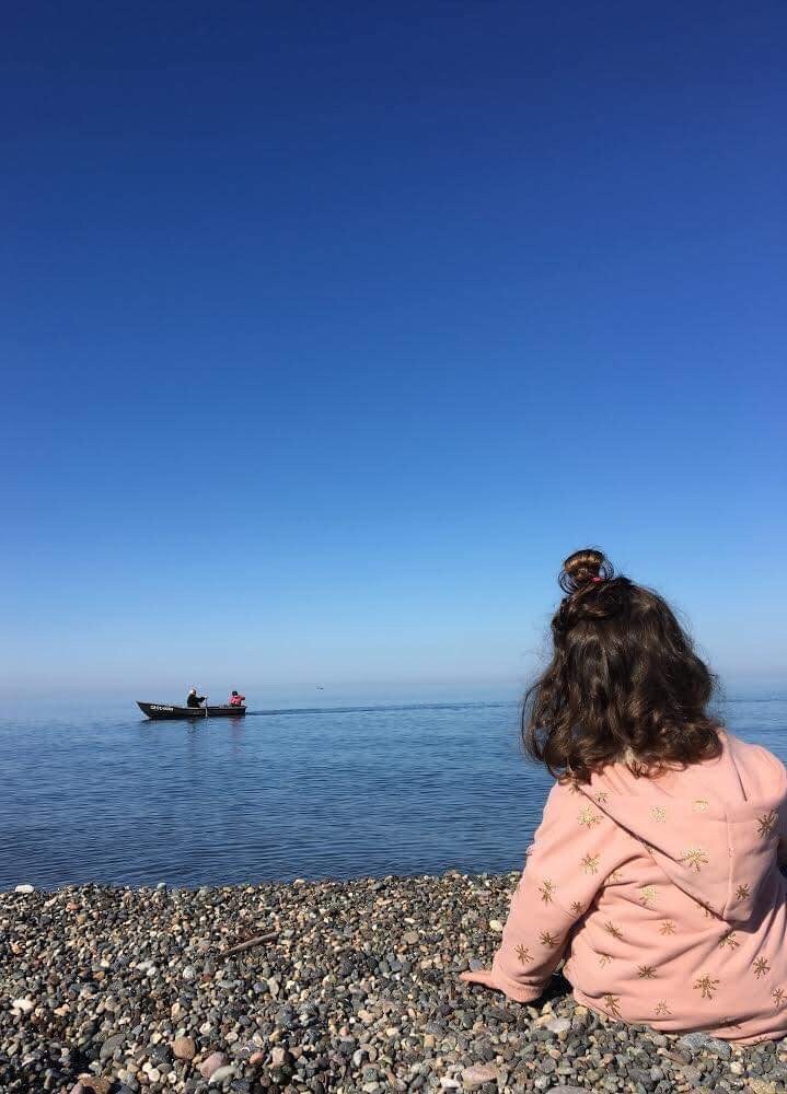 A child is seated on a pebbly shore, facing the calm blue water. A small boat is seen in the distance with two individuals aboard. The sky above is clear and bright, contributing to the serene coastal atmosphere.