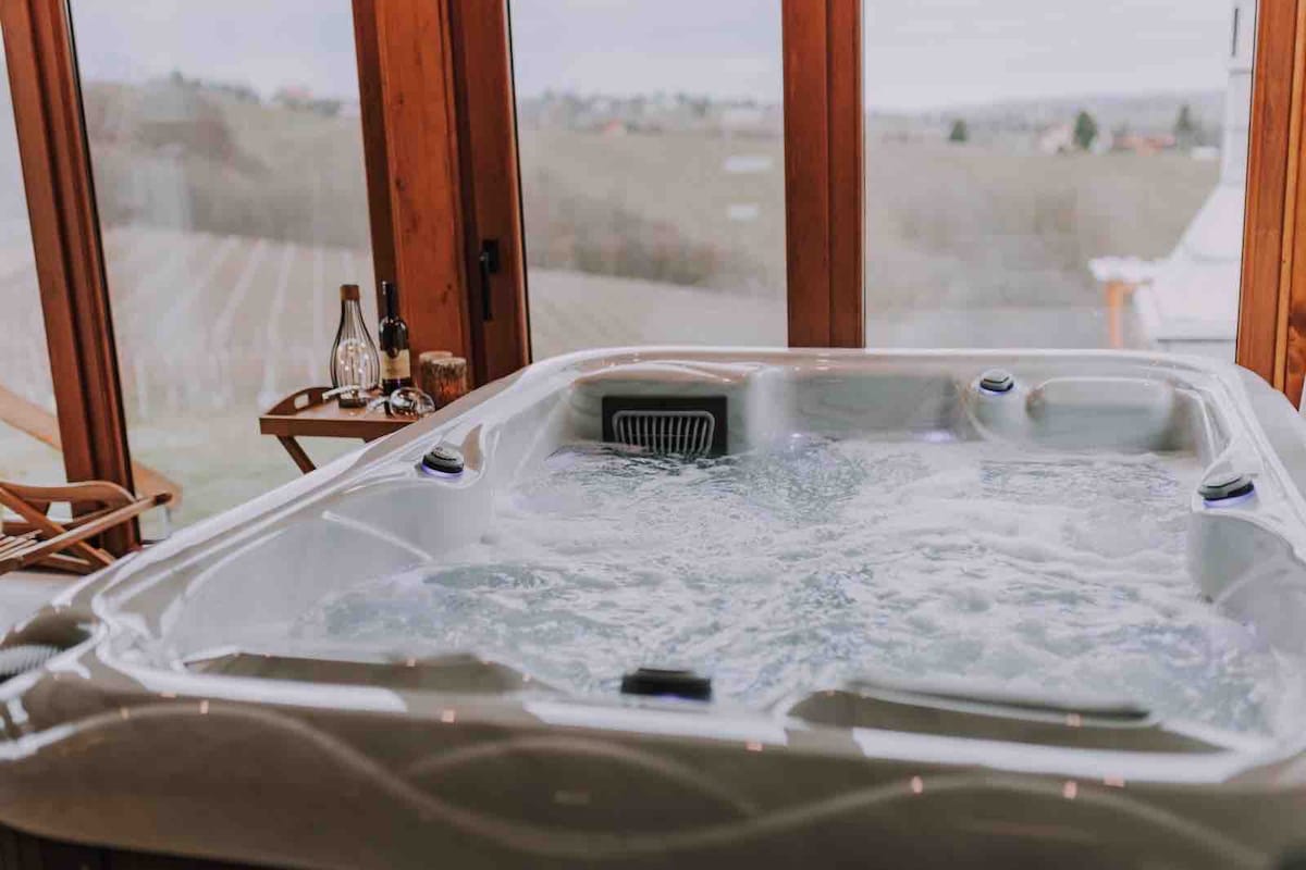 A relaxing hot tub is positioned by large windows, providing views of the surrounding vineyards and countryside. Wooden details are present in the room, complemented by small tables featuring bottles nearby. Natural light filters in through the windows, enhancing the serene atmosphere.