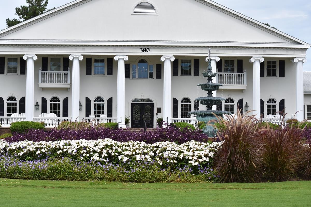 A stately white building features prominent columns and an elegant entrance. The well-manicured lawn is adorned with colorful flower beds, showcasing bright white and purple blooms. A decorative fountain stands in the center, enhancing the serene ambiance of the setting.