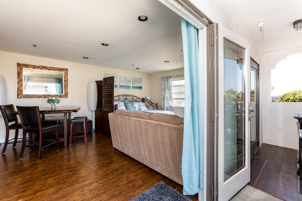 The interior of a modern beach studio is displayed, featuring a cozy sitting area with a comfortable sofa and a dining table for four. Light wood flooring complements the bright blue accents, while large windows allow natural light to fill the space. A private balcony door opens to scenic views.