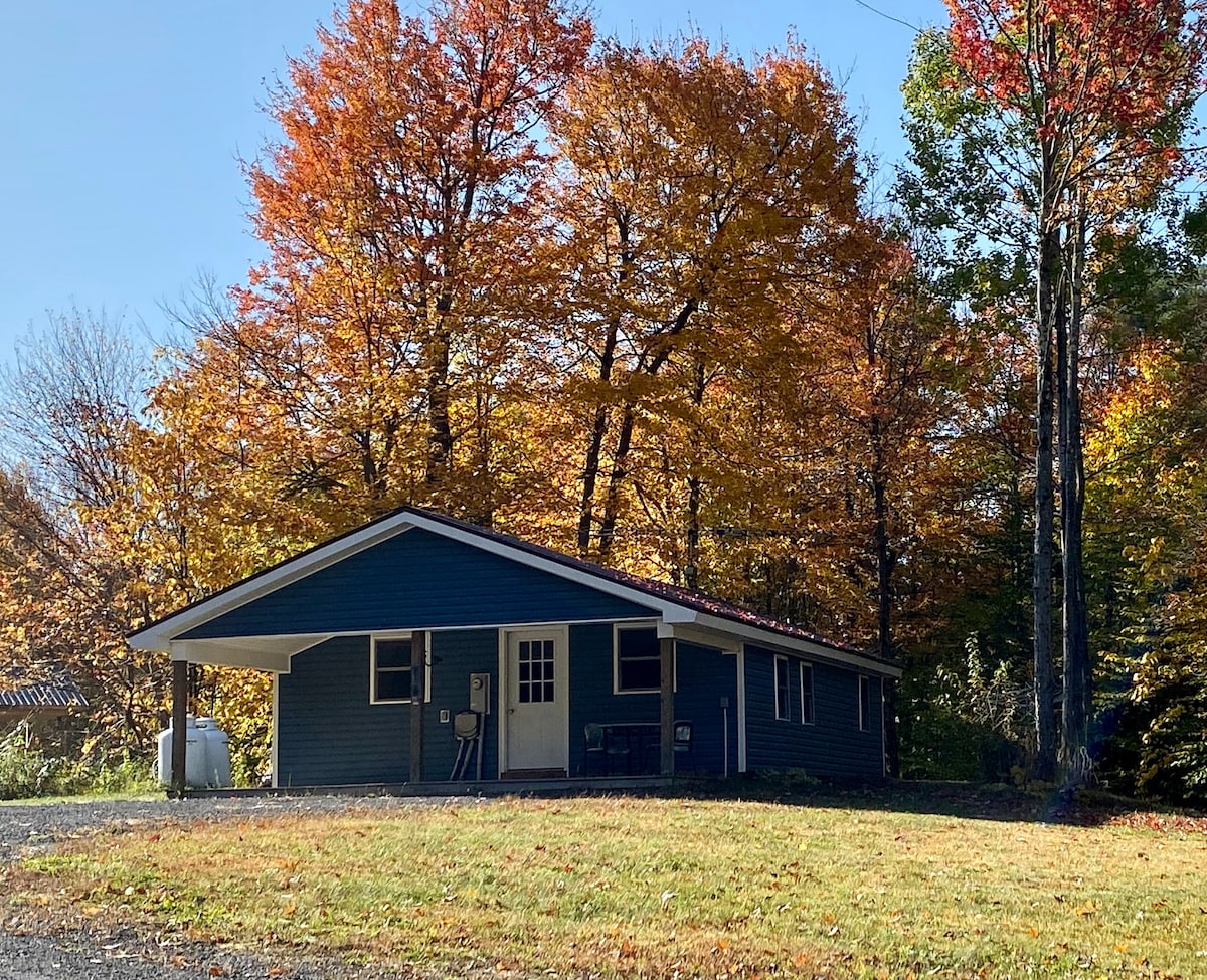 A private house is set against a backdrop of vibrant autumn foliage. The structure features a covered porch and large windows, with a gravel driveway leading to the entrance. Surrounding trees offer a natural, peaceful setting.