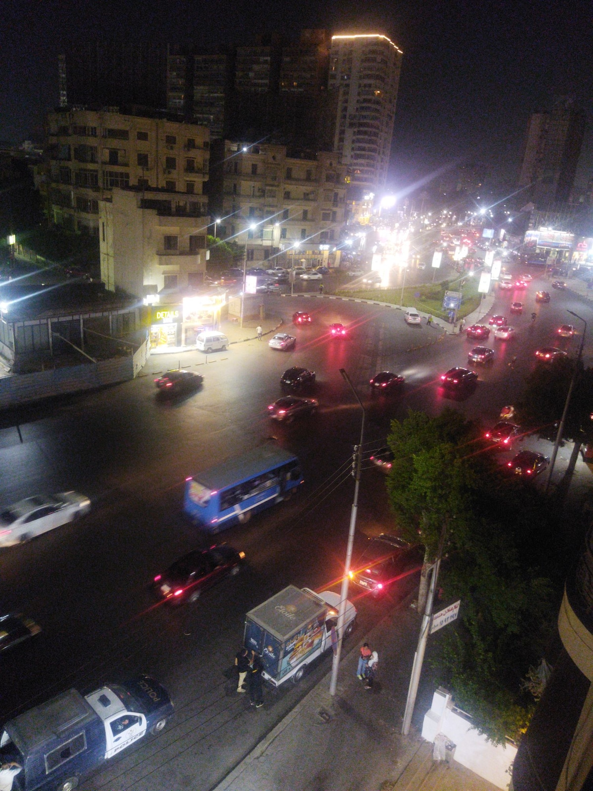 A bustling urban scene is captured at night, showcasing a busy intersection illuminated by streetlights and colorful signs. Various vehicles, including cars and buses, are in motion on the road, while buildings line the street, contributing to the lively atmosphere.