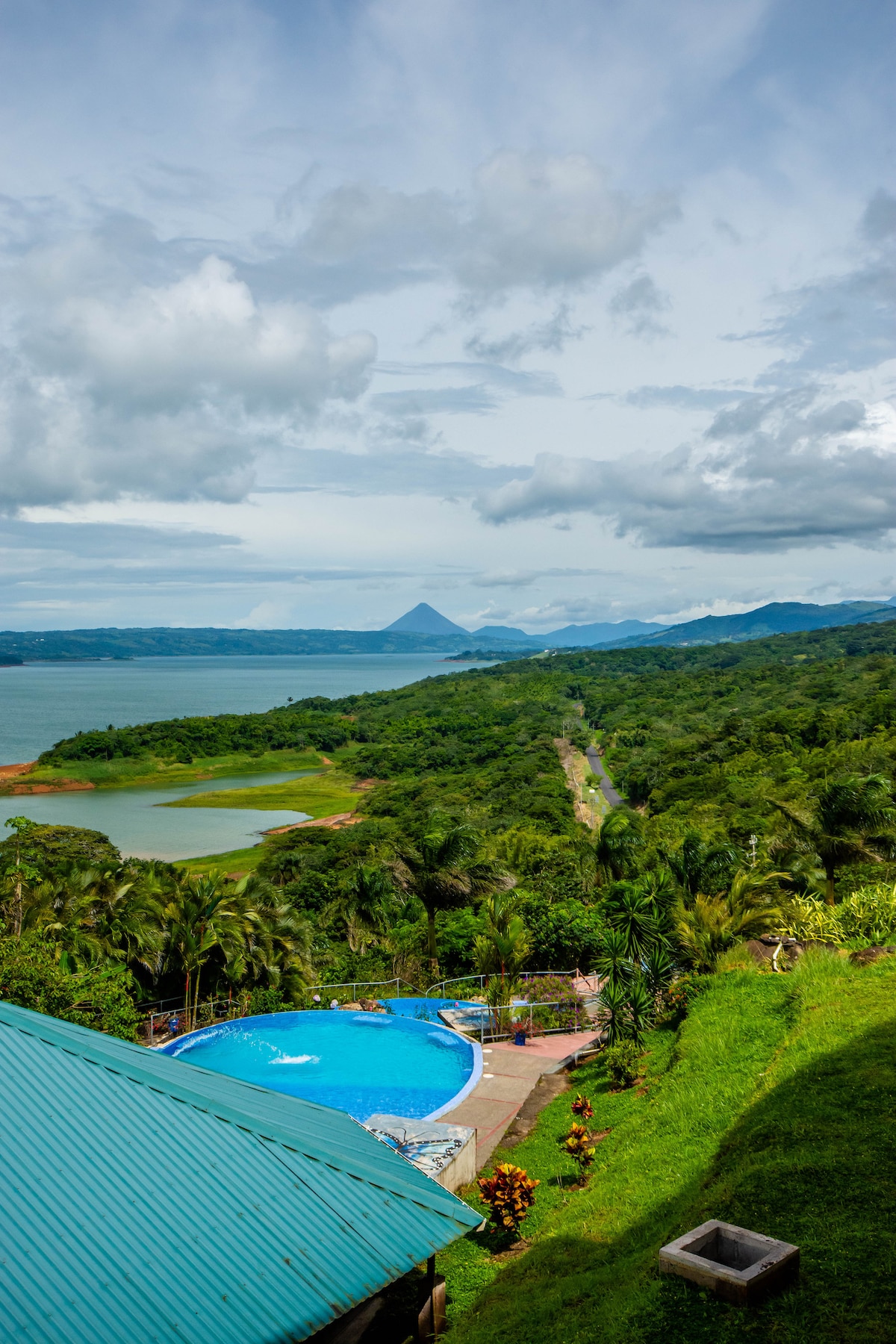 A panoramic view captures a vibrant blue pool surrounded by lush greenery. In the background, Lake Arenal extends toward distant mountains under a partly cloudy sky, showcasing the natural beauty of the landscape.