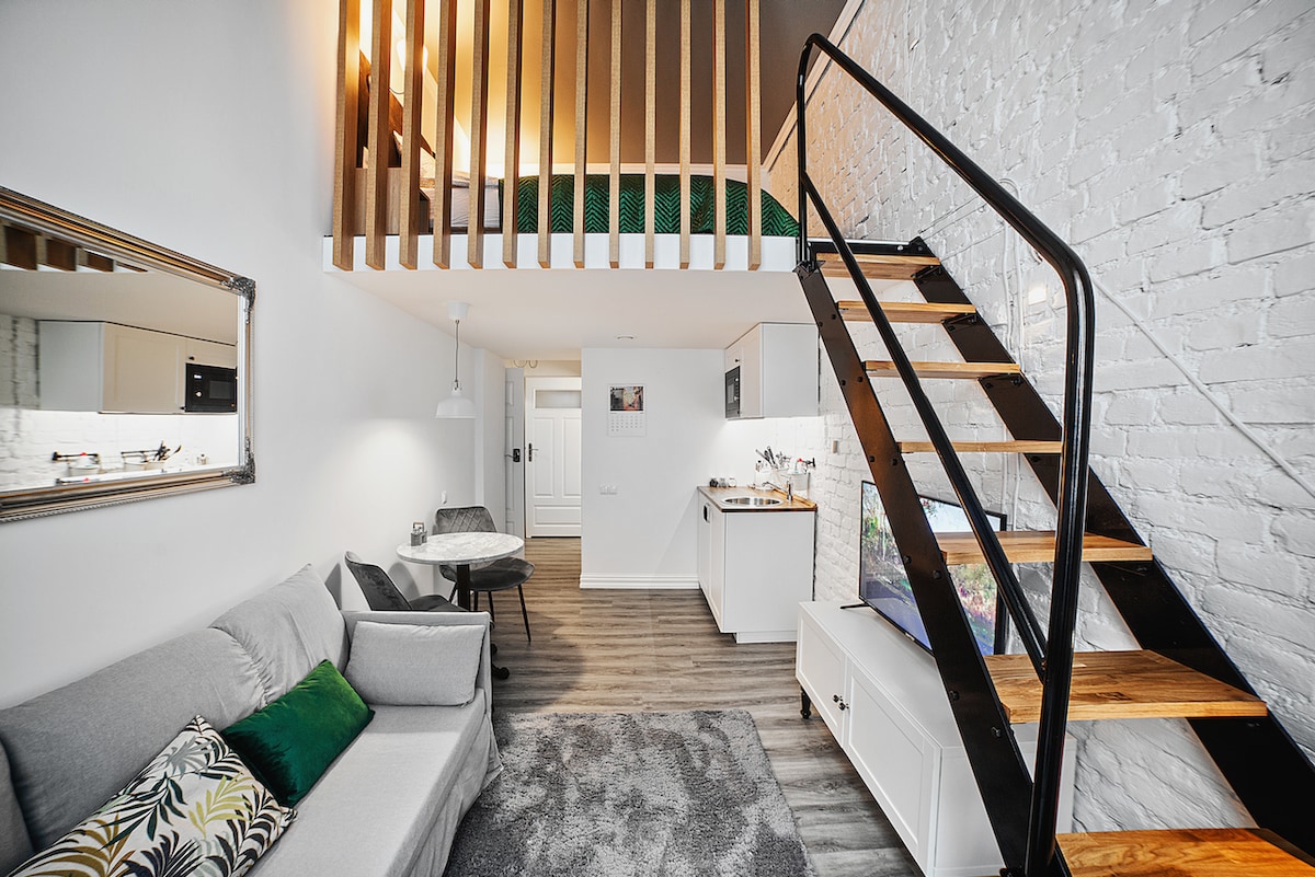 A bright and airy living area is highlighted by a comfortable sofa, dining table, and a staircase leading to a lofted sleeping area. The modern kitchen is visible, featuring sleek cabinetry and essential appliances, while large windows allow natural light to enhance the space.