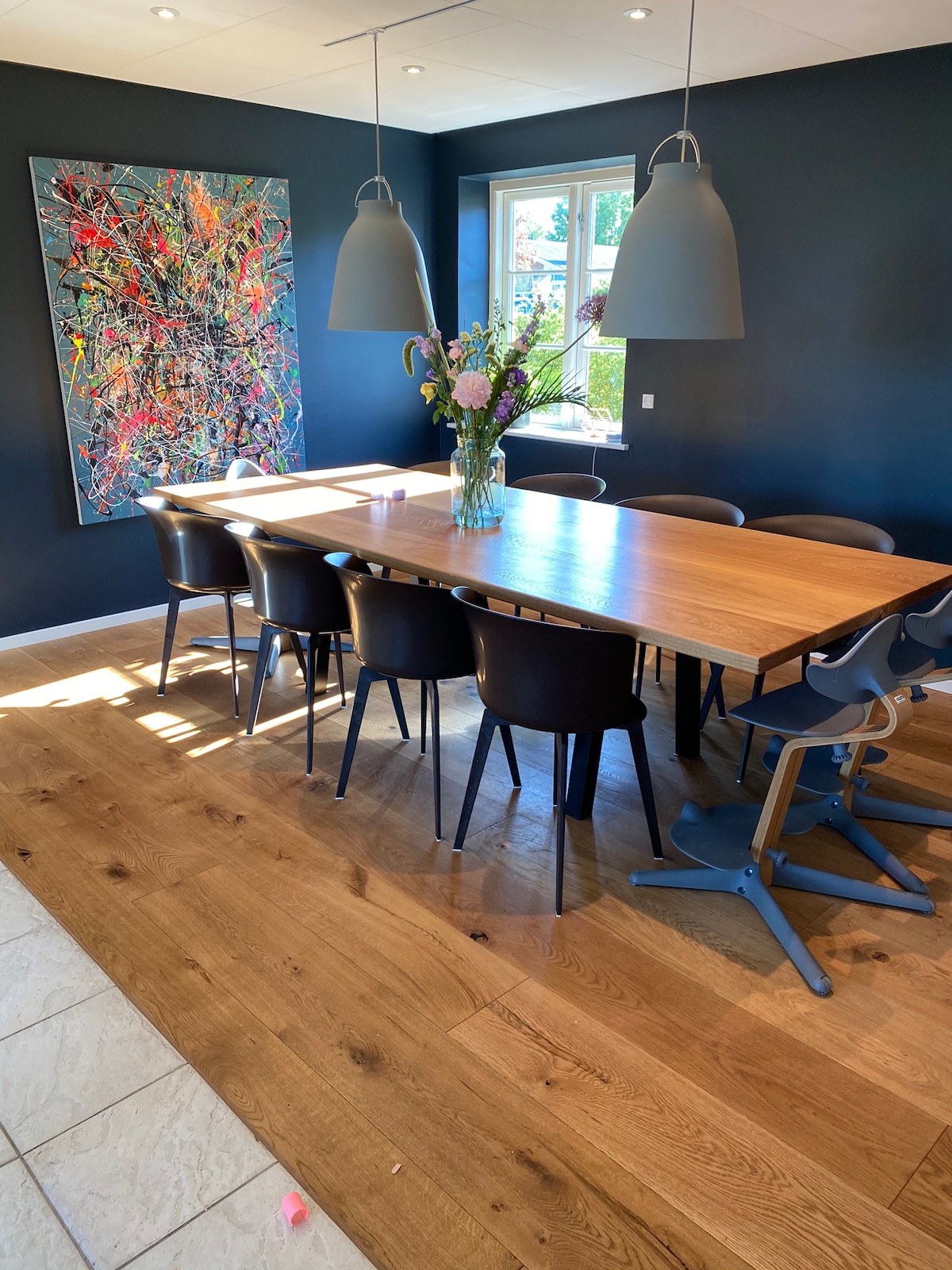 A spacious dining area features a large wooden table surrounded by black chairs, illuminated by natural light from a window. Two pendant lights hang above the table, complementing a colorful abstract art piece on the dark blue wall.