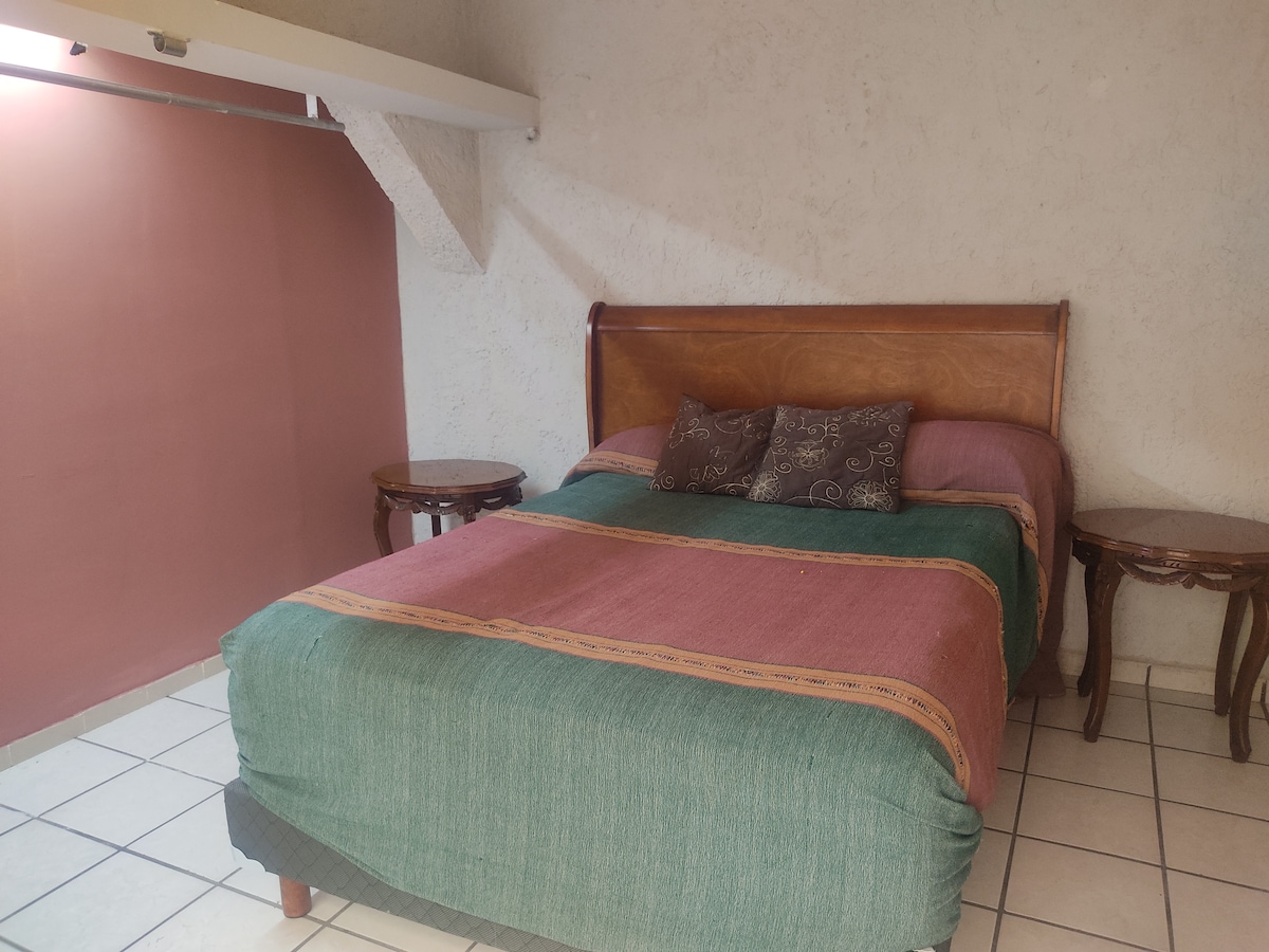 A sleeping area features a spacious bed with a multicolored blanket and decorative pillows. Two small wooden side tables flank the bed. Light from a nearby window enhances the warm tones of the walls, creating a comfortable and cozy environment.