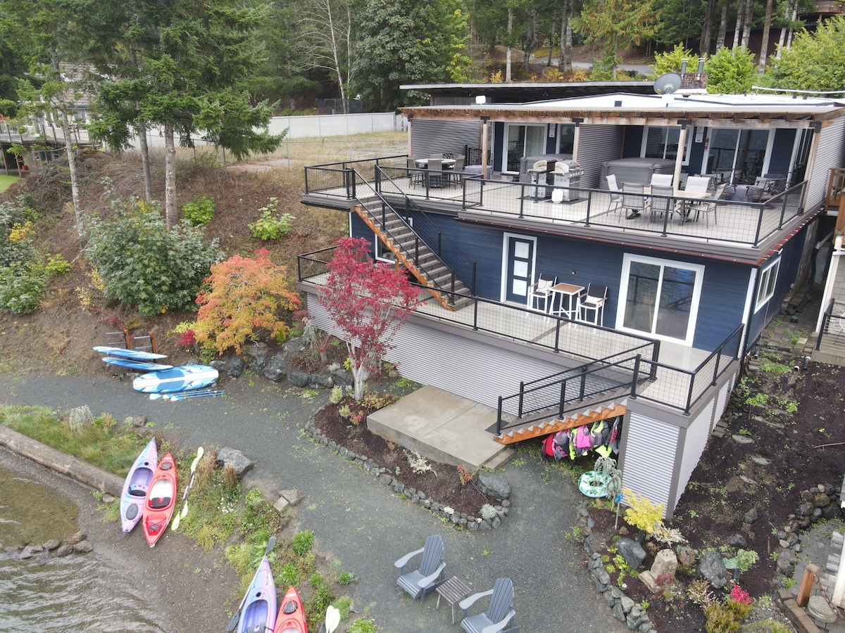 A multi-level duplex sits adjacent to the lake, featuring a large deck with seating. Colorful kayaks and paddleboards are positioned on the shore, while lounge chairs are arranged near the water's edge. Natural greenery and seasonal plants enhance the setting.
