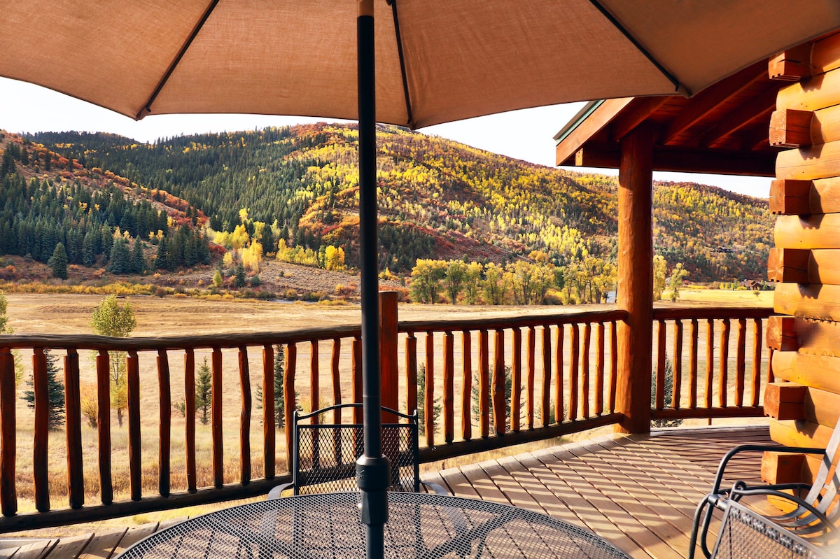 A wraparound deck offers seating with a shaded table and views of rolling hills adorned with autumn foliage. The landscape is complemented by a grassy meadow, surrounded by trees, under a clear sky.