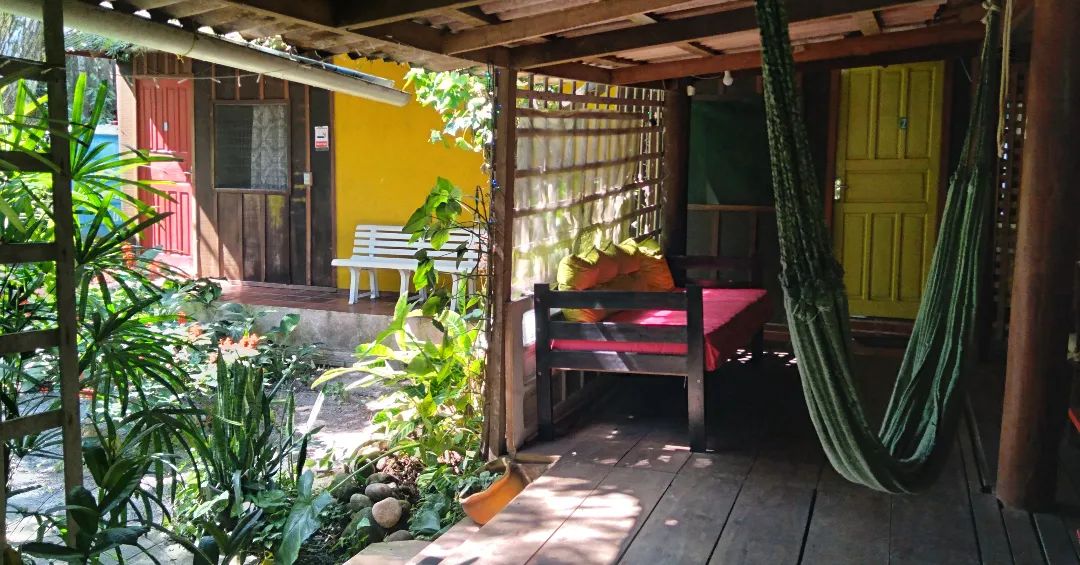 A covered patio area reveals a vibrant yellow door leading to an interior space. A welcoming seating arrangement includes a large bed draped in colorful bedding and a hammock, surrounded by lush greenery and decorative plants.