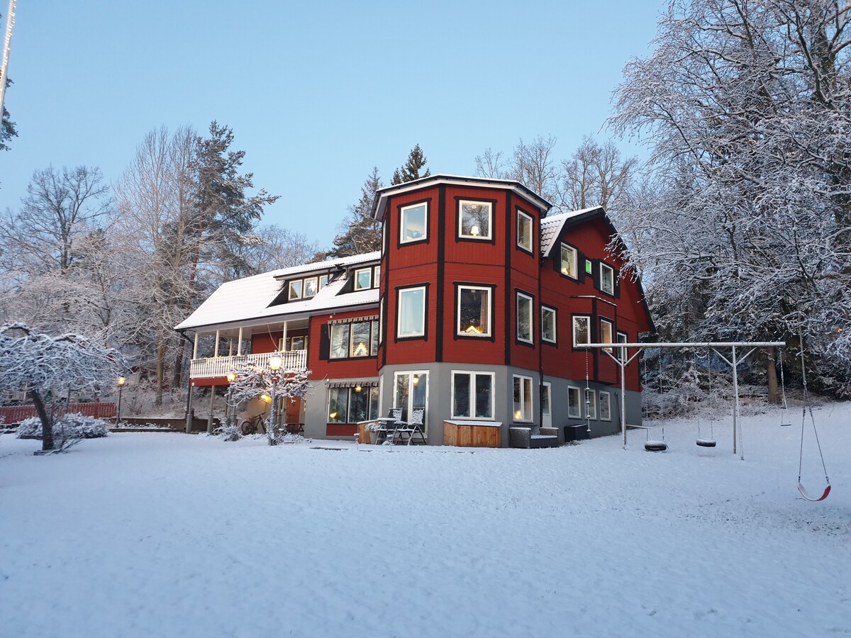 A spacious red house with multiple windows sits on a snow-covered yard, surrounded by trees. The building features a unique architectural design, with several levels and angles. A swing set is visible in the yard, adding to the outdoor recreational space.