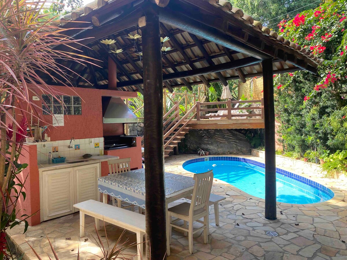 An outdoor area presents a pool surrounded by natural stone paving. A shaded space features a wooden table with accompanying chairs, adjacent to a grill and a kitchen area. Lush greenery and flowering plants create a serene environment.