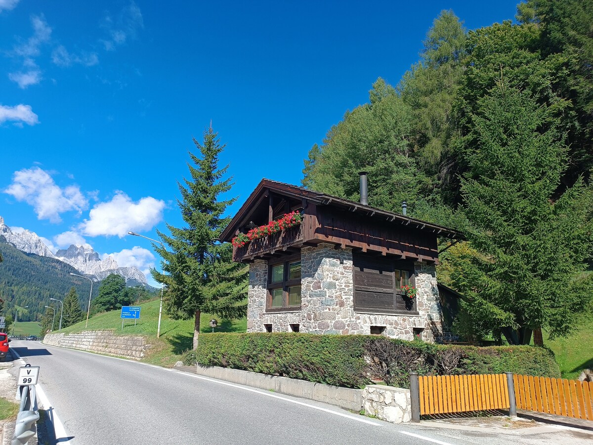 A charming mountain cottage showcases a rustic stone facade complemented by wooden beams and window boxes filled with greenery. Lush trees surround the property, and the scenic mountain landscape forms a stunning backdrop against a clear blue sky.