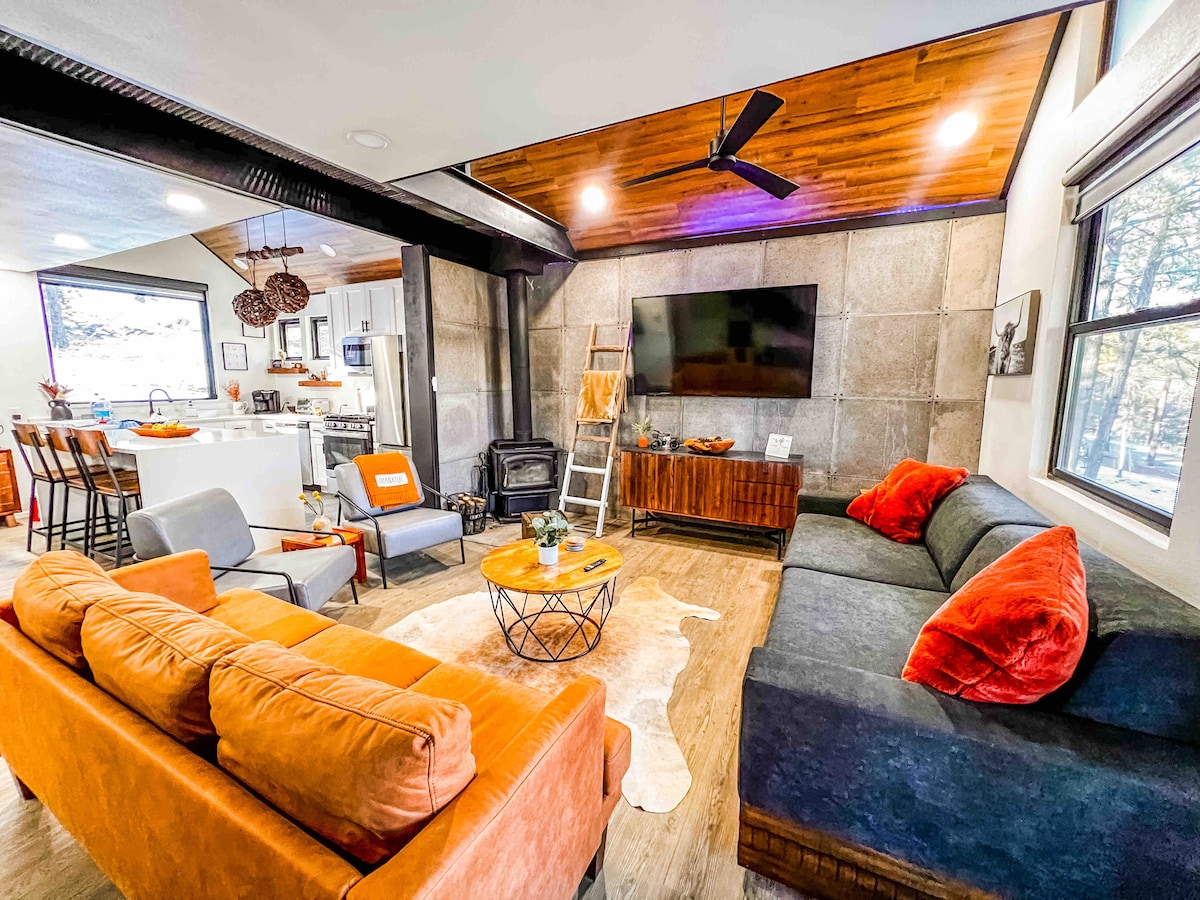 The living area features a combination of a dark sectional and a bright orange sofa, arranged around a circular coffee table on a light rug. A 60-inch flat-screen TV is mounted on a textured wall, with a cozy wood stove nearby. Natural light filters in from windows leading to the kitchen.