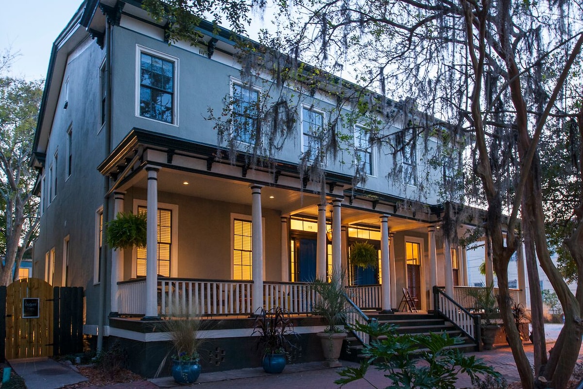 The historic townhouse features a spacious front porch, supported by tall columns, with potted plants adding greenery. Warm light glows from the windows, creating an inviting entrance. Spanish moss hangs from nearby trees, enhancing the southern charm of the property.