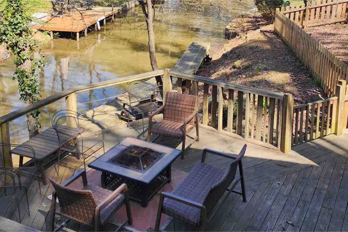 A wooden deck area features a fire pit surrounded by four wicker chairs. The deck overlooks the water, with a boat dock visible nearby. The setting is framed by trees and a fenced backyard, offering a secluded outdoor space.