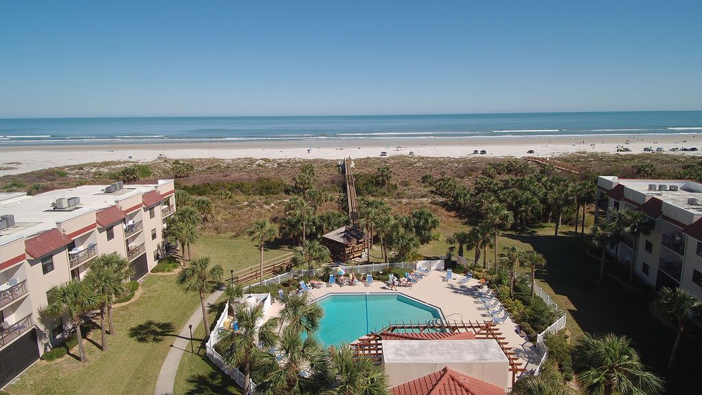 An aerial view captures a serene beachfront setting, showcasing a sparkling pool surrounded by lush green palm trees and lounge chairs. The expansive beach stretches along the horizon, with gentle waves lapping at the shore under a clear blue sky.
