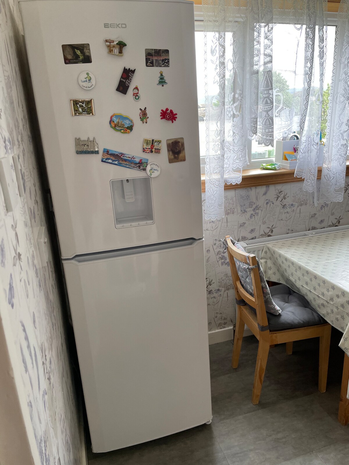 A white refrigerator stands against the wall, decorated with various magnets. A small dining table, adorned with a patterned tablecloth, is situated nearby. Natural light filters through lace curtains, illuminating the kitchen space while floral wallpaper adds a touch of charm.