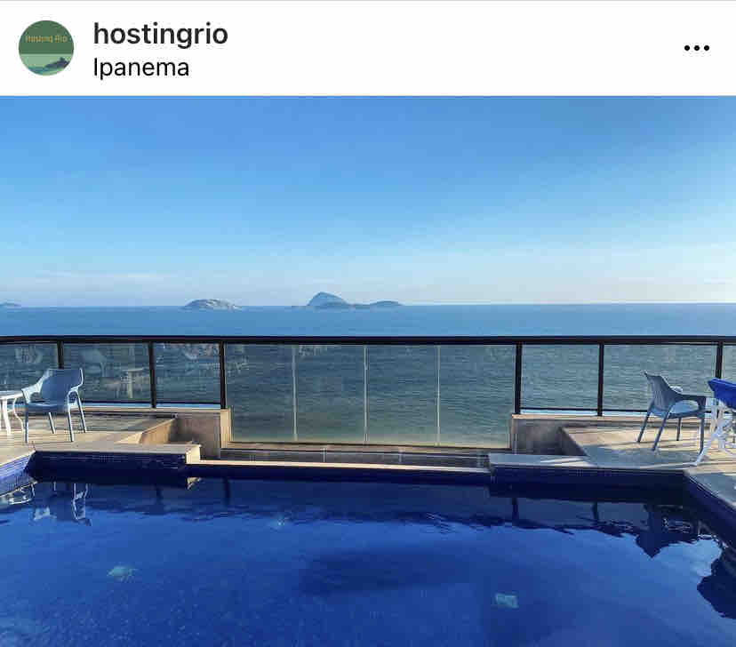 A rooftop pool offers a tranquil blue surface reflecting the sky, with a glass railing providing an unobstructed view of the ocean and distant islands. Several white lounge chairs are positioned around the pool area, creating an inviting space for relaxation.