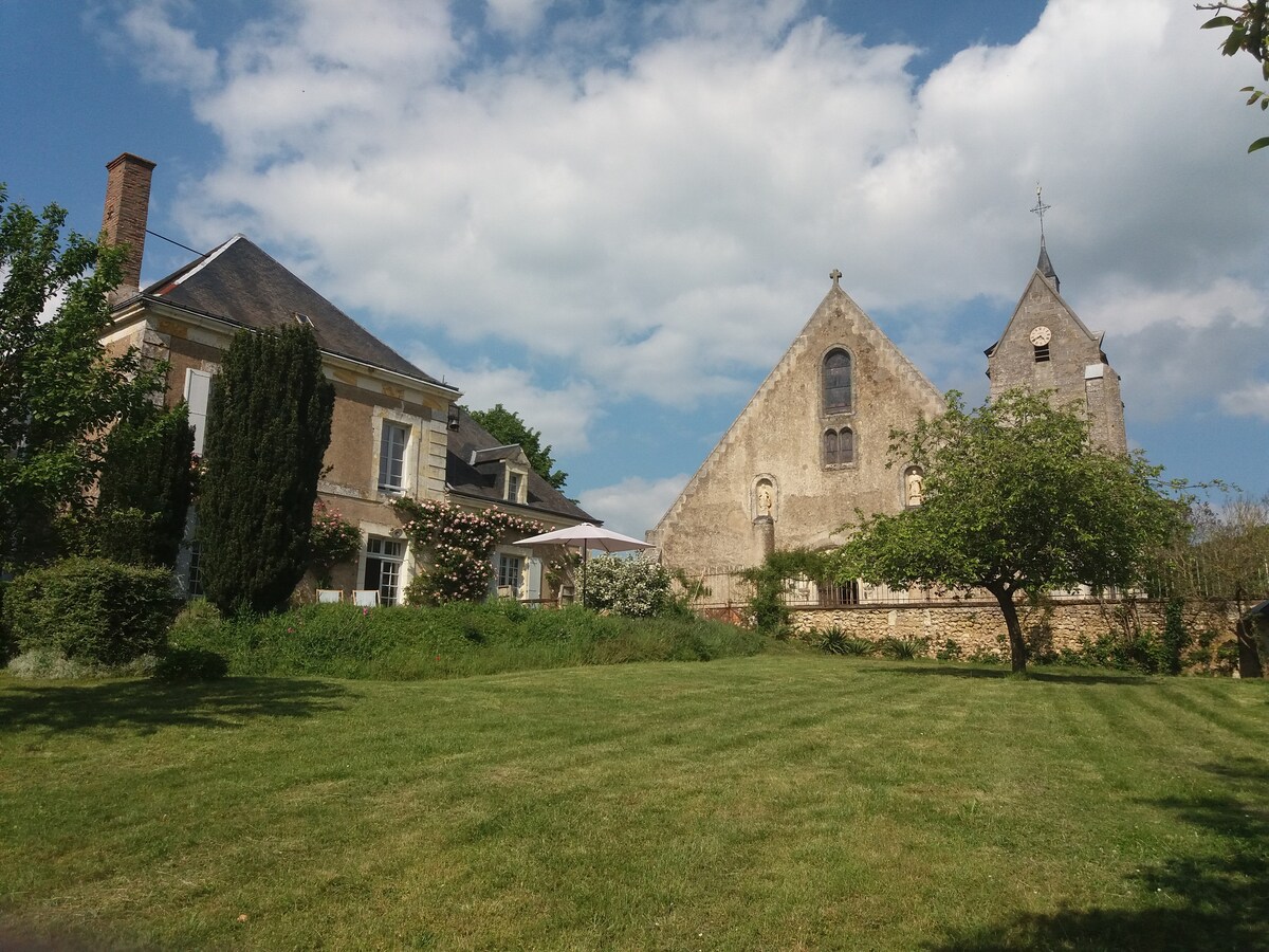 A charming house is situated adjacent to a historic church, both surrounded by a landscaped garden. A lush green lawn extends across the foreground, while light clouds drift across the sky, enhancing the serene setting.