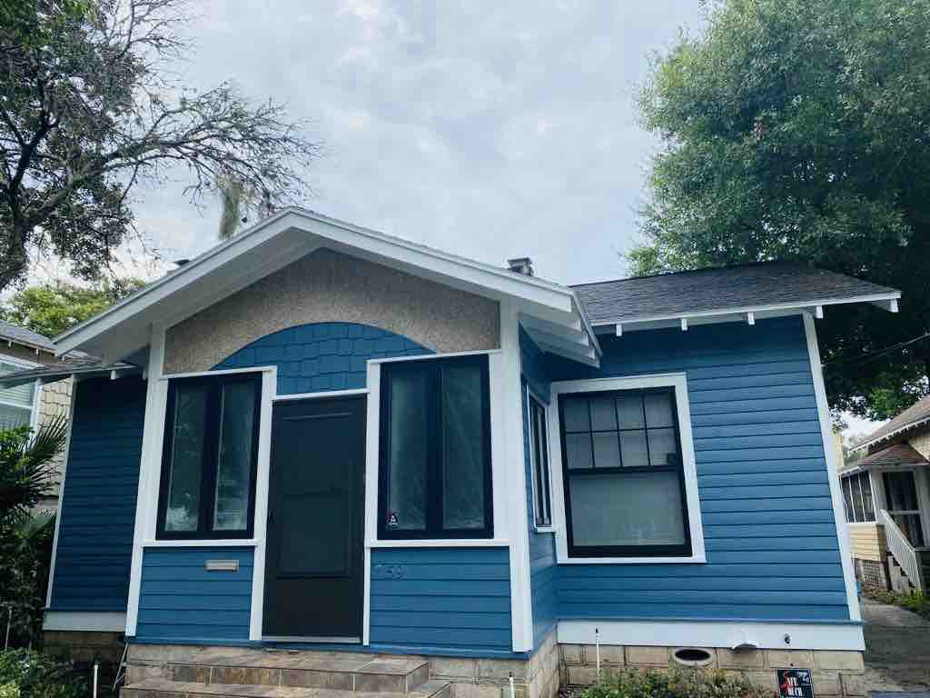 The exterior of a charming blue house is displayed under a partly cloudy sky. The entrance features a large black door framed by two windows. The architectural style showcases clean lines and a welcoming façade, with greenery surrounding the property.