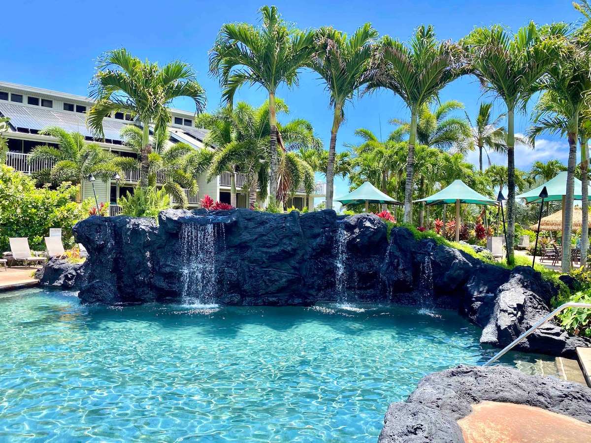 A swimming pool features a naturalistic rock waterfall surrounded by vibrant tropical landscaping. Tall palm trees and green umbrellas provide shade around the pool area, while lounge chairs are positioned nearby for relaxation. The clear blue water invites guests to enjoy a refreshing dip.