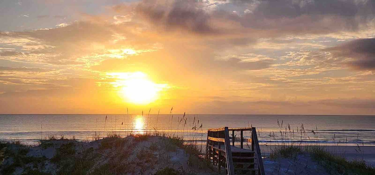 A beautiful oceanfront sunset is captured, with rich hues of orange and gold reflecting on the water. Grass and sandy dunes are visible in the foreground, while a wooden walkway leads toward the beach, inviting exploration of the serene coastal environment.
