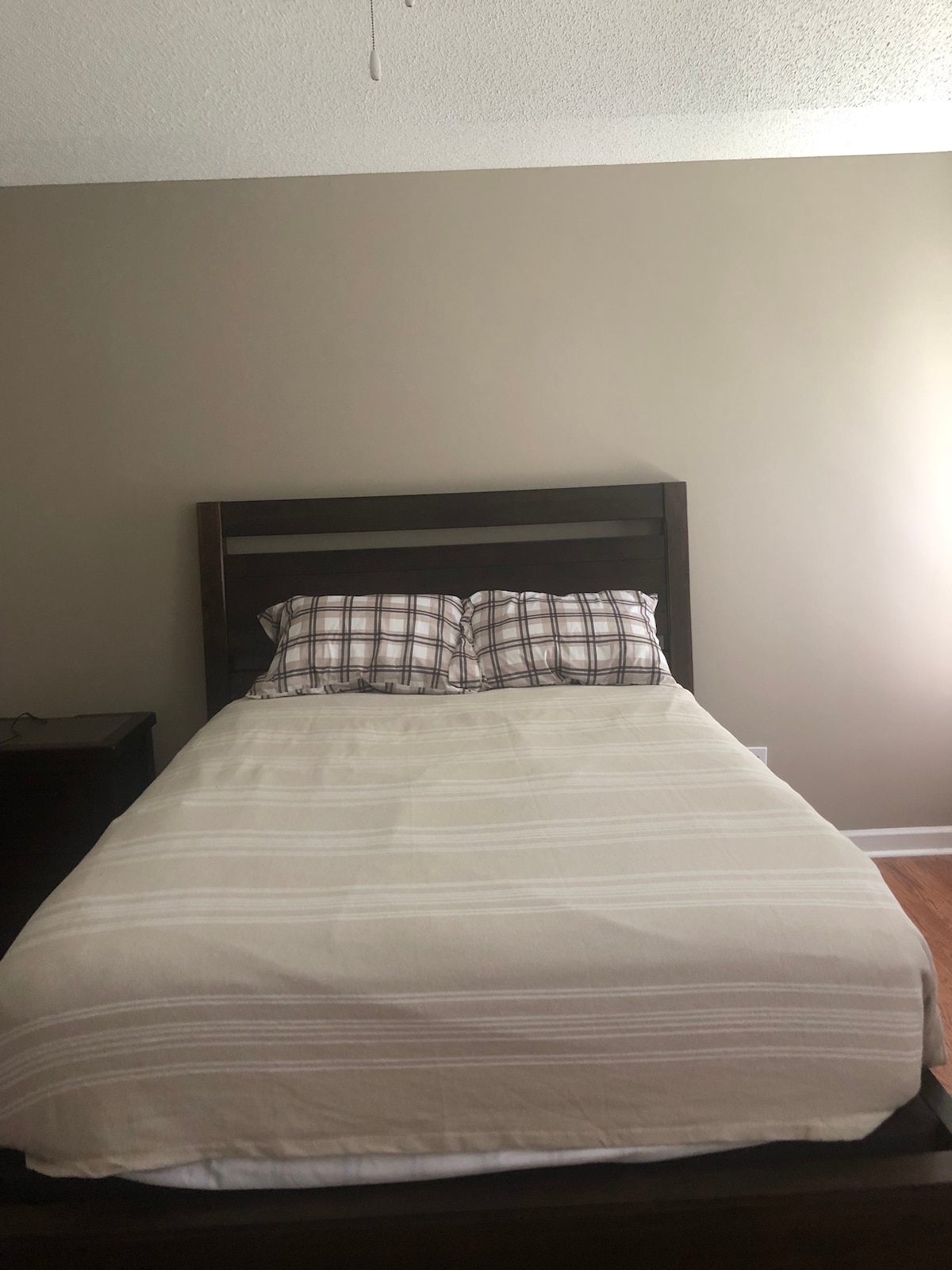 A cozy bedroom features a neatly made bed with a light-colored striped blanket and plaid pillows. A dark wooden headboard complements the light beige walls, while natural light enters from a nearby window, creating a calm and inviting appearance.