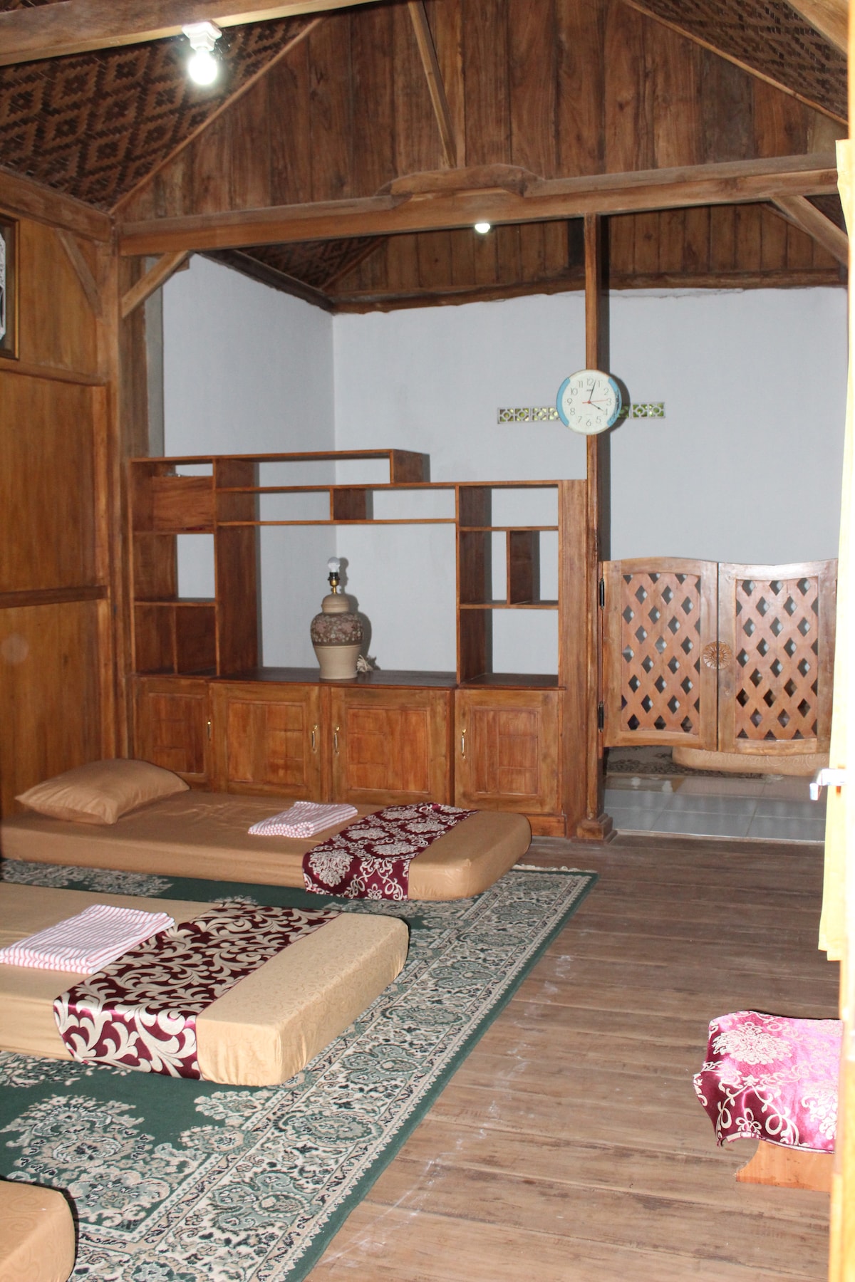 A simple interior showcases wooden accents and a warm atmosphere. Eight floor mattresses are placed on a patterned rug with additional textiles. A wooden shelving unit and decorative elements are positioned against the wall, alongside a clock and a privacy screen.