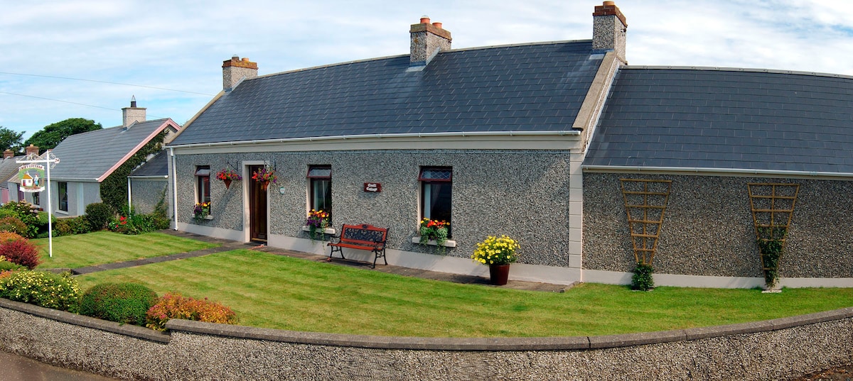 The exterior of the cottage showcases a charming stone façade, complemented by a slate roof. A well-maintained garden with colorful plants and flowers borders a pathway leading to the entrance, where a wooden bench offers a welcoming spot to relax.