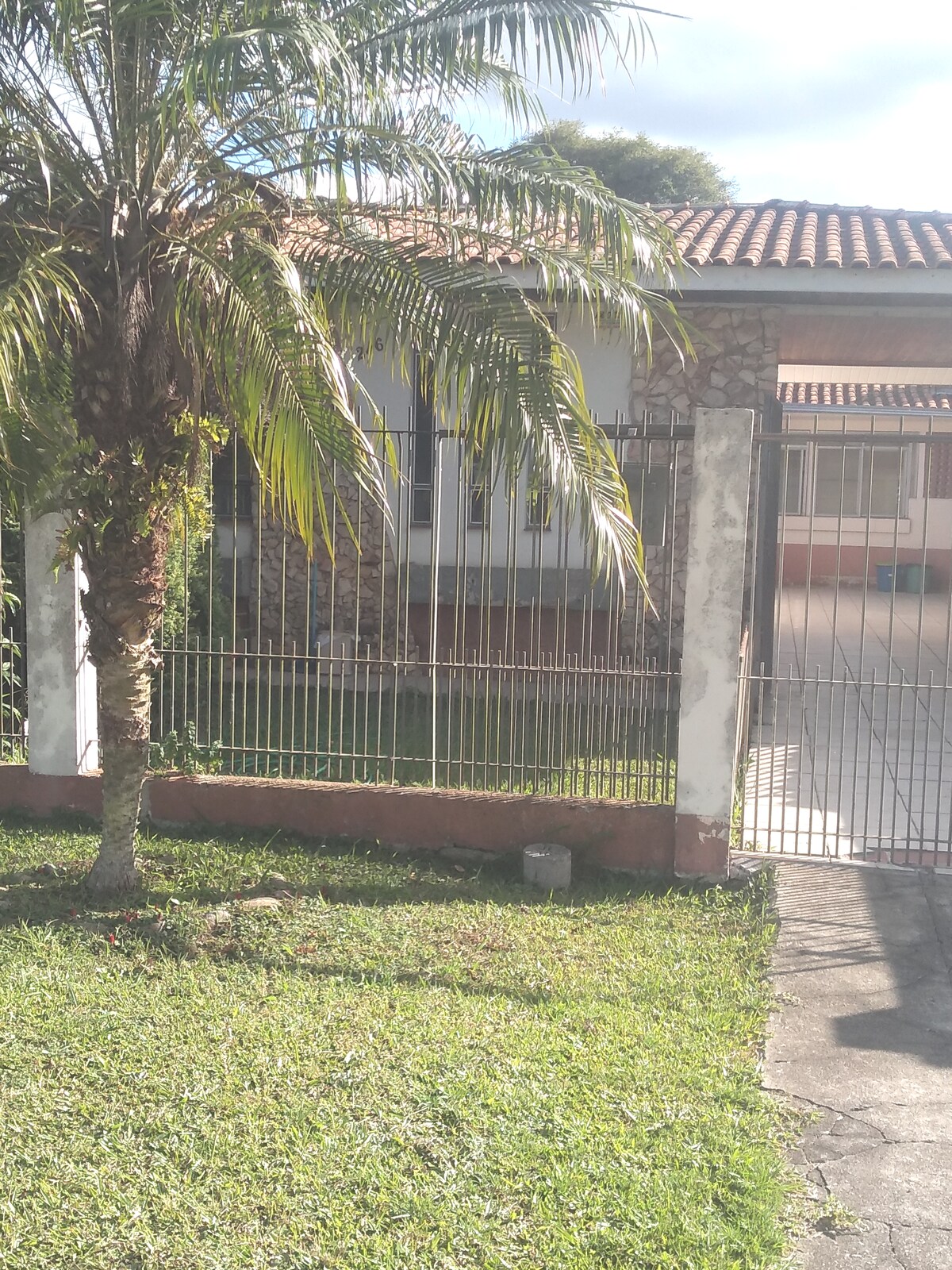 A well-maintained exterior features a stone facade and a tiled roof. A green lawn is bordered by a metal fence, with a palm tree positioned near the entrance. Clear skies provide natural lighting to the inviting entrance of the property.