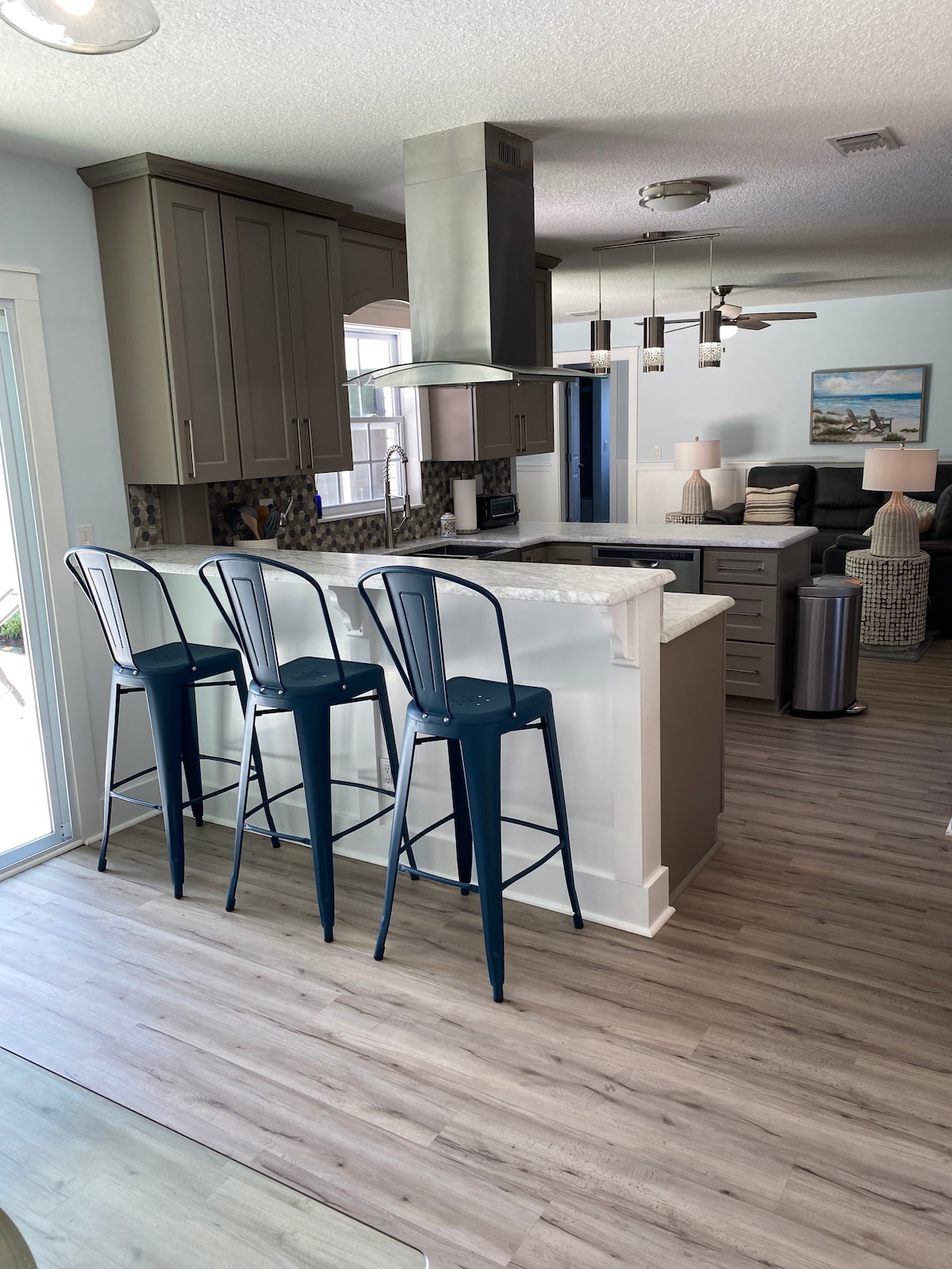 The kitchen features modern cabinetry and a white countertop, with three blue metal bar stools positioned at the island. A stainless steel hood and appliances complement the open layout, which transitions to a cozy living area furnished with a sectional sofa.