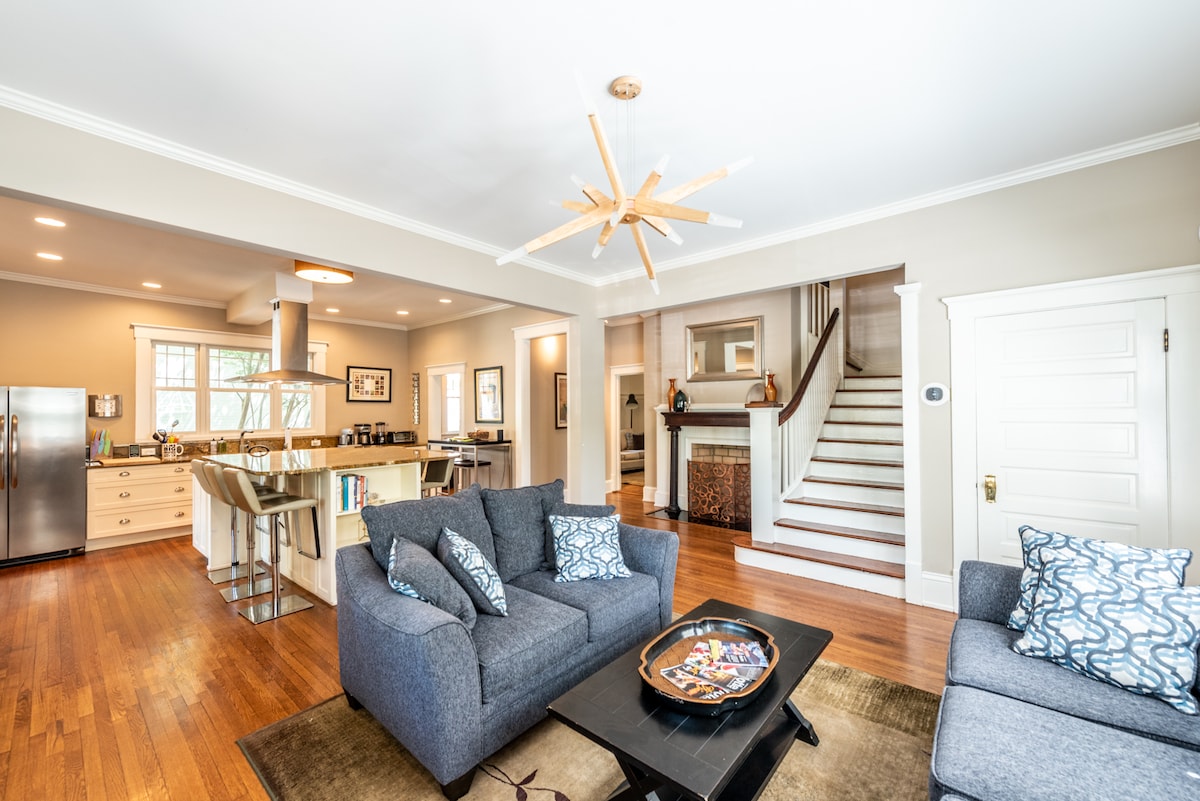 An open living space features a cozy seating area with gray sofas, accented by decorative pillows. A modern kitchen with stainless steel appliances and a large island is visible, alongside a dining area. Light wood flooring enhances the natural light from surrounding windows.