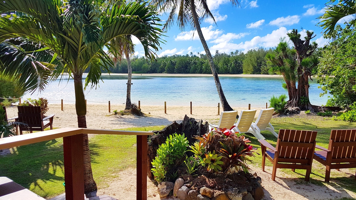 A tranquil beachfront view showcases a serene lagoon, framed by palm trees and lush greenery. Several lounge chairs are positioned on the sandy shore, inviting relaxation. The clear blue sky reflects on the water, enhancing the peaceful atmosphere of the outdoor space.