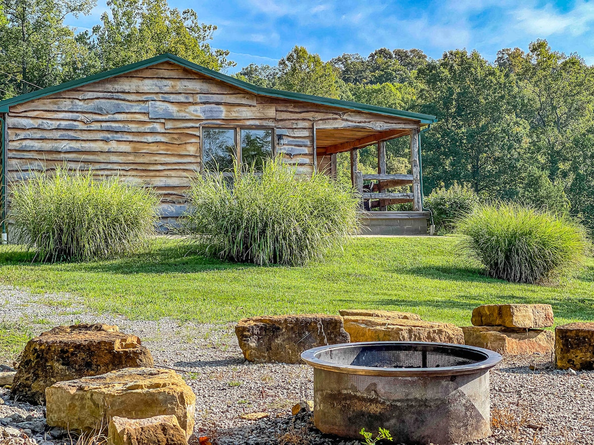 The cabin features a rustic wooden exterior with large windows reflecting the surrounding greenery. A fire ring is positioned nearby, surrounded by natural stone seating, offering a cozy gathering space. Lush grass and ornamental plants enhance the outdoor setting.