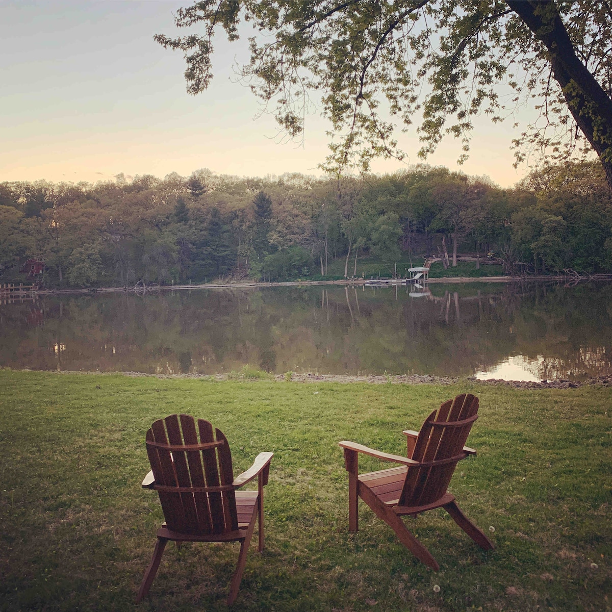 Two wooden Adirondack chairs are positioned on a grassy area along the water's edge, facing a calm river. Reflections of trees and the surrounding landscape are visible in the still water, creating a serene outdoor setting.