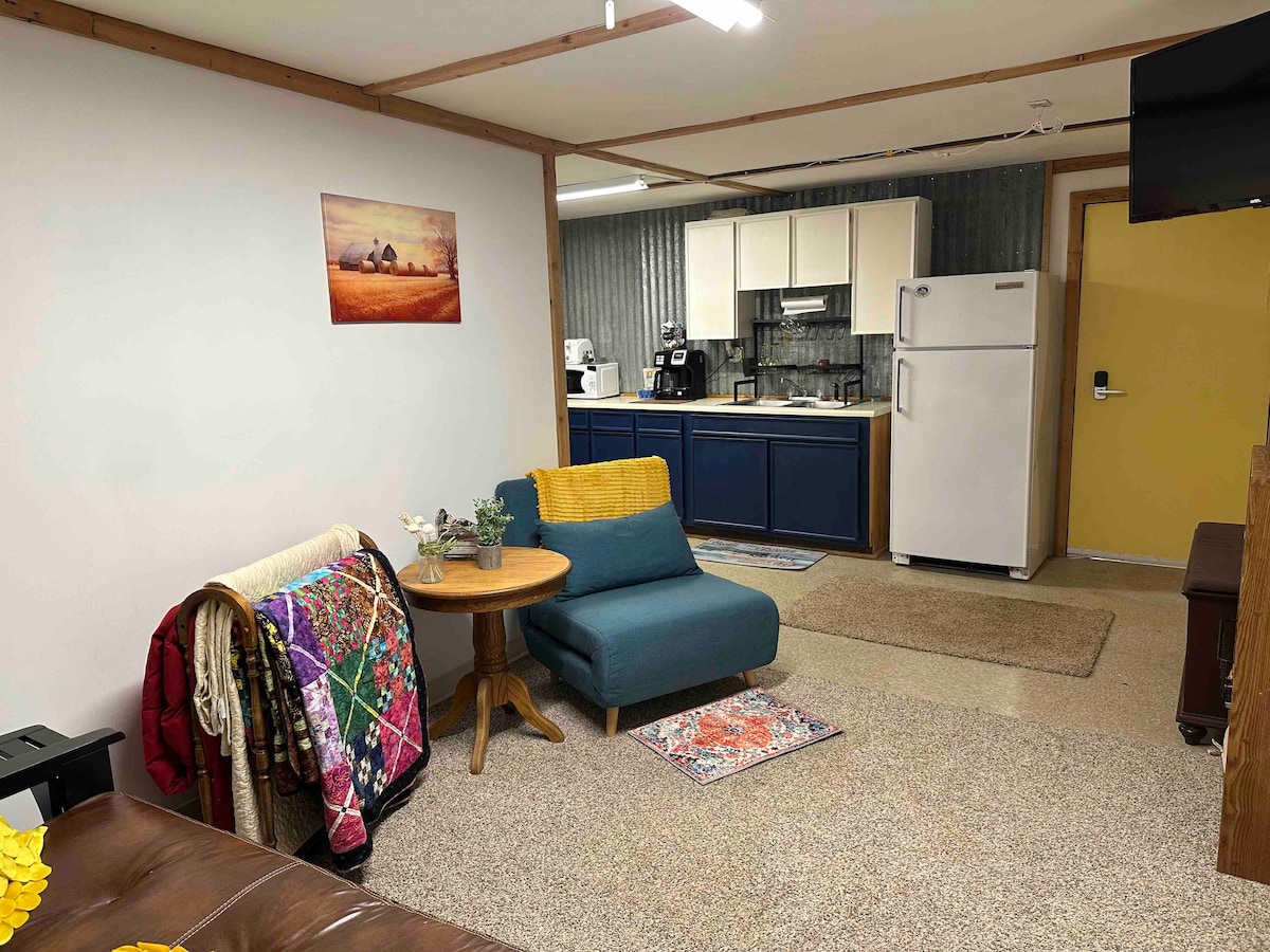 The living area features a cozy seating arrangement with a blue chair and small wooden table. A colorful quilt drapes over the sofa. The kitchen, equipped with essential appliances, is visible in the background, highlighted by navy cabinetry and warm lighting.