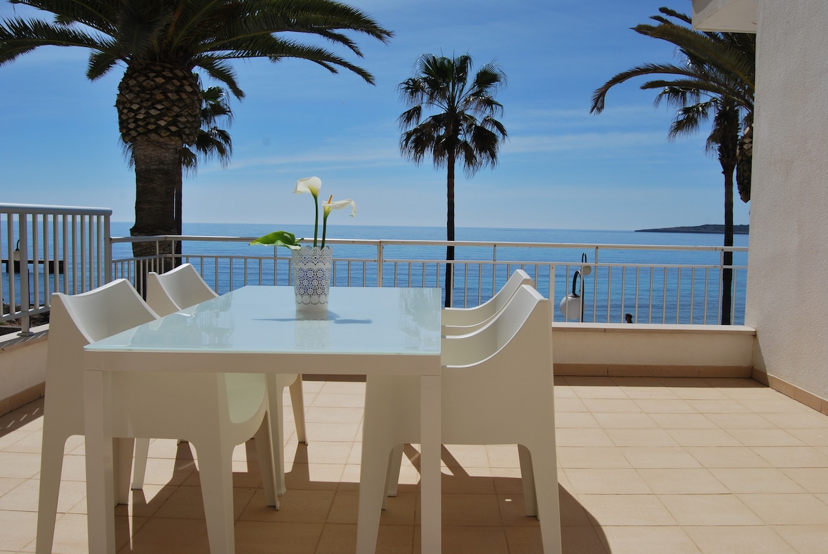 A private terrace is featured with a white dining table and four chairs, offering views of the Mediterranean Sea. Two palm trees are visible in the background, and a vase with fresh flowers is placed at the center of the table.