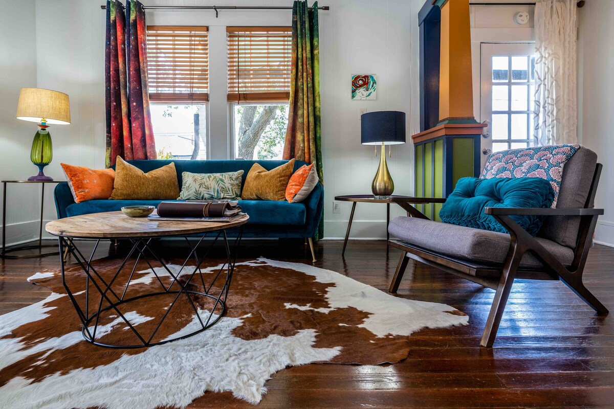 A cozy living area features a dark blue sofa adorned with colorful pillows, accompanied by a comfortable armchair. A round coffee table rests on a patterned area rug, with warm light emanating from a nearby floor lamp and a side lamp, creating a welcoming ambiance.