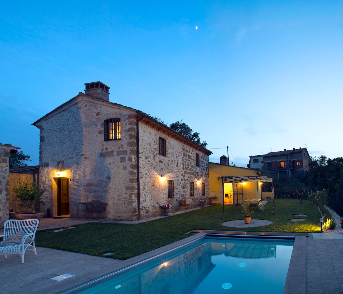 A charming stone house is shown under a evening sky, with warm lights glowing from its windows. The well-maintained garden features a refreshing salt-water pool and patio area, surrounded by lush greenery and a gazebo.