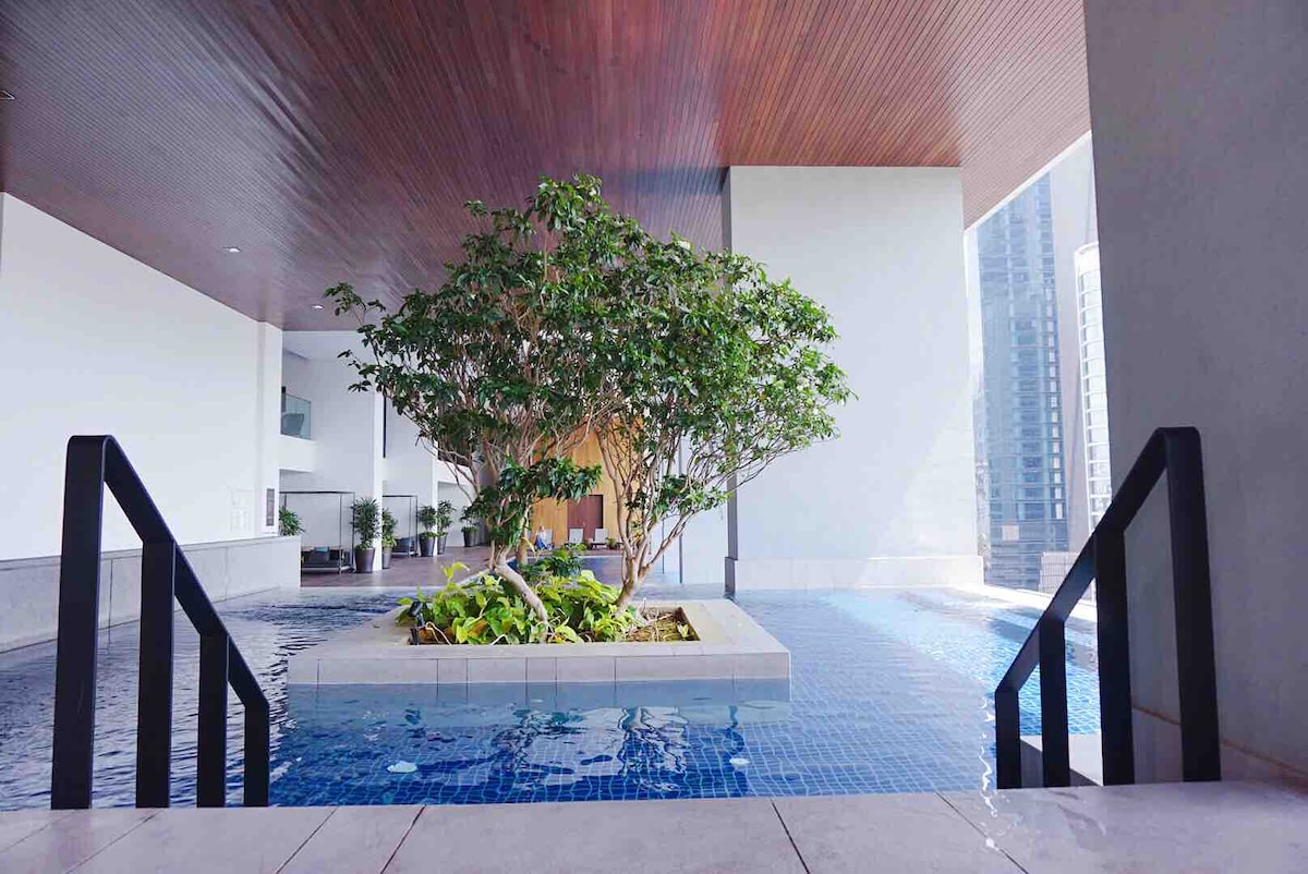 A serene pool area is depicted, featuring a central planter with lush greenery under natural light. The pool's edge is outlined by sleek stairs leading in, while the surrounding architecture offers a modern aesthetic with clean lines.