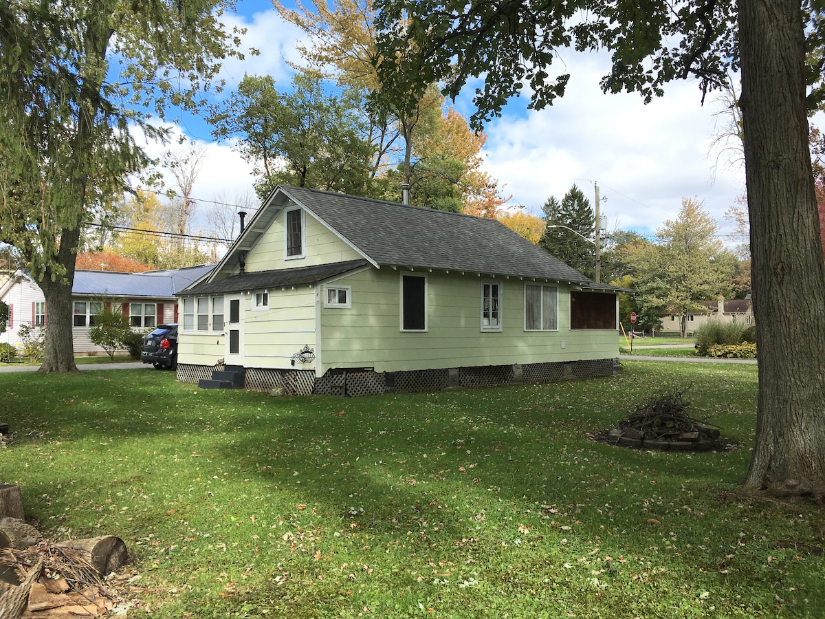The cottage is set back on a grassy yard, surrounded by mature trees. A light green exterior is visible, featuring multiple windows. The structure has a gabled roof and a covered porch, with a view of the quiet neighborhood and pathway leading to the street.