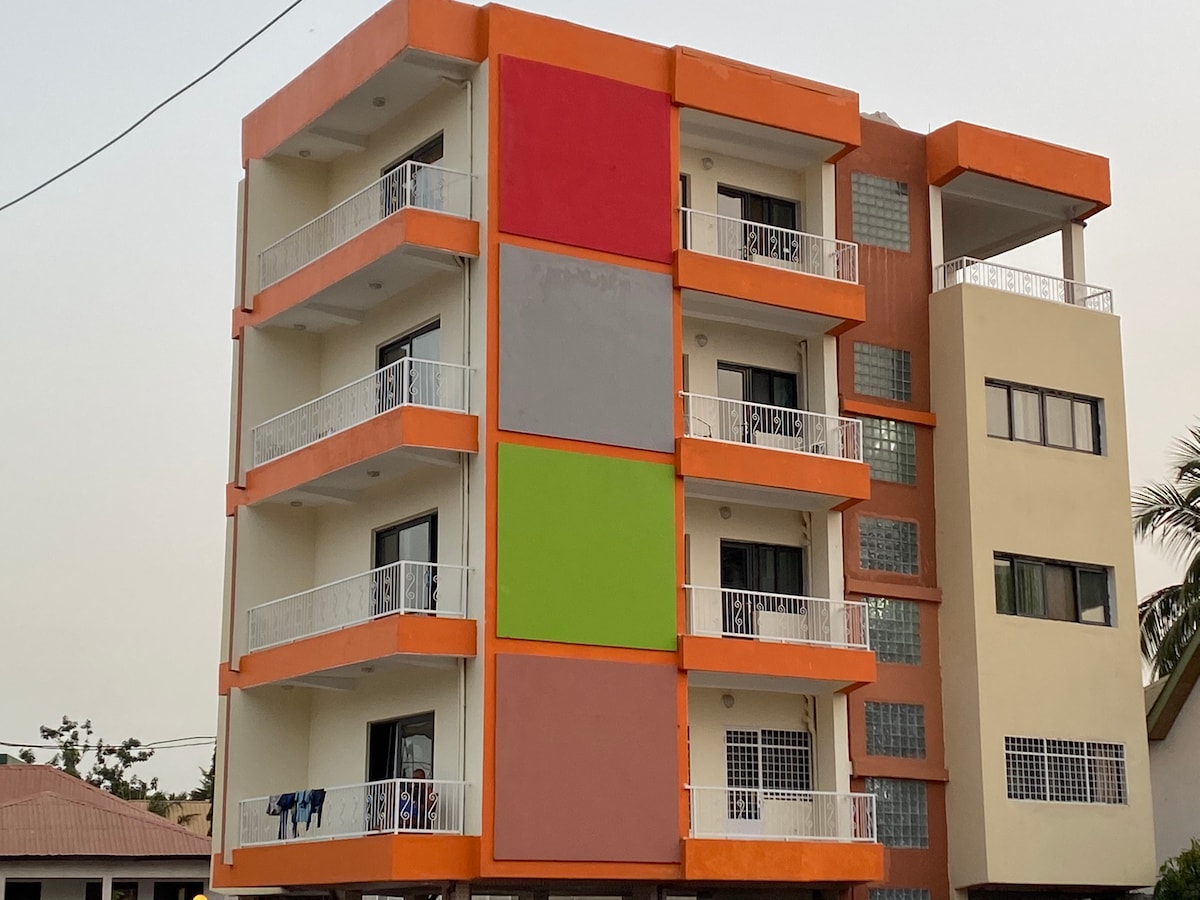 A modern building featuring four levels with balconies framed by white railings. The facade showcases colorful sections in red, orange, gray, and green, creating a vibrant appearance. Adjacent to it, another building presents a more muted exterior.